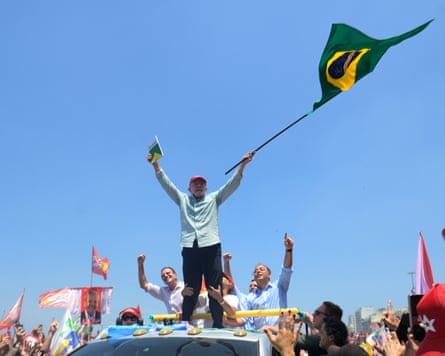Lula da Silva stands on a car roof holding a book and flag at a rally.