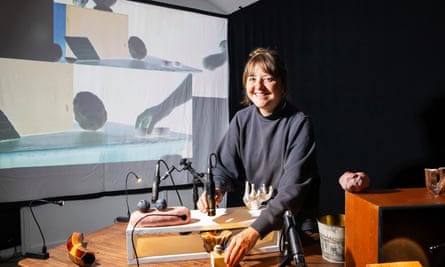 A woman shows her sculptures in front of a projected screen of hands holding objects.
