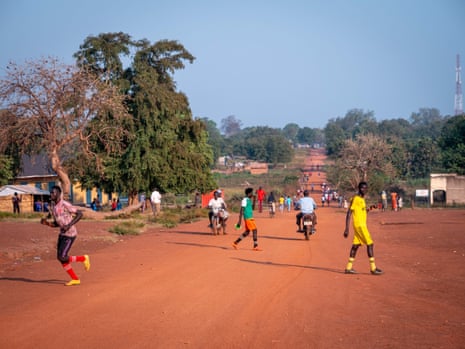 People walking along a wide road of deep orange-red dirt with huts on either side and motorbikes passing