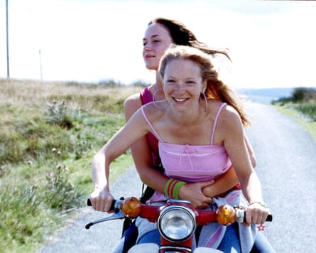 A young woman holds her arms around another young woman, both in vests and with long hair, as they drive on a moped down a country lane