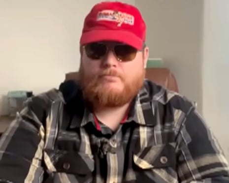 Joshua Bonehill-Paine wearing sunglasses and a red cap sits indoors in a plaid shirt