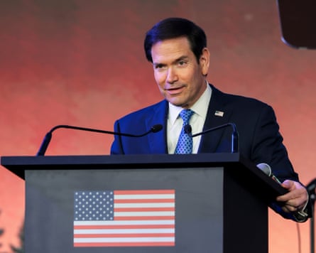 Marco Rubio speaks at a podium bearing a US flag, with microphones nearby
