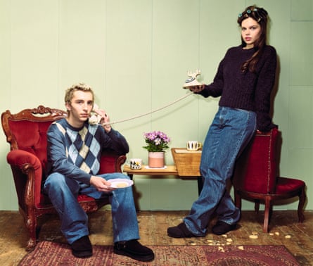 Young man and woman looking like old people, him sitting in a red armchair, holding the receiver of a vintage phone whose base she is holding