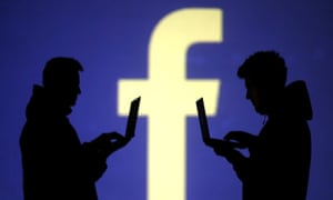 Silhouettes of laptop users are seen next to a screen projection of Facebook logo