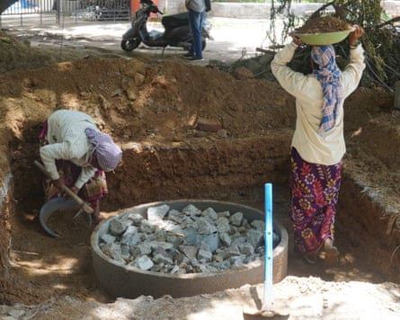 Two Indians in lungi-type wraps, one filling a rubber basin with soil in a square hole while another carries a full basin of soil on their head