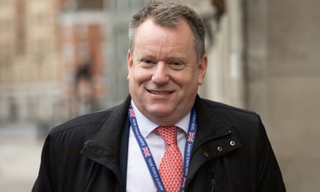 Lord Frost smiling in the street, in an ill-fitting dark coat, red tie and blue lanyard