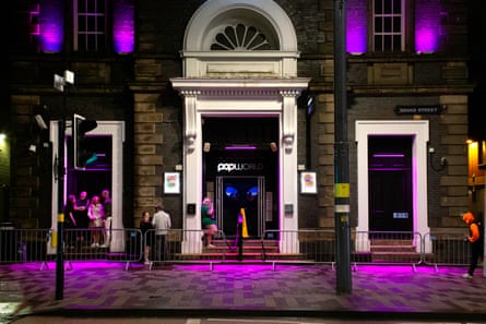 An imposing door framed by a white wooden arch and pillars, and pik neon lights. A few people are on the street.