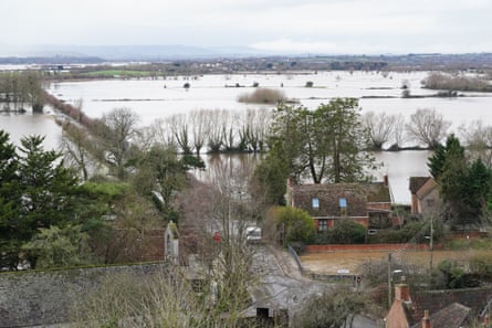 Flood water surrounds rural cottages and their outbuildings, and covers the fields behind them like a large lake. The landscape is flat with a few small trees above the water.