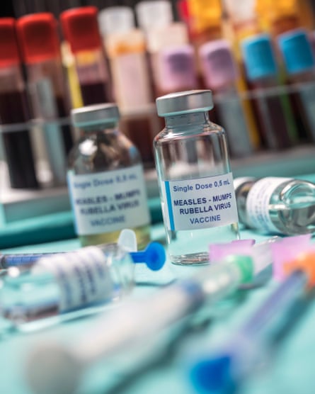 Different vials of triple vaccine against measles, mumps and rubella (MMR) seen against backdrop of colourful test tubes and vials with syringes in foreground