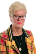 Helen Pitcher, portrait photo: she is in her 60s with short blond hair and wears black-rimmed glasses, a black top, a bright yellow, red and black tartan jacket and gold necklace plus red lipstick