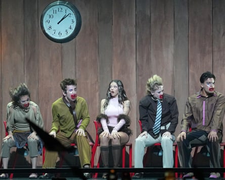 A young woman dressed in pink, sitting on a chair in a wood-panelled room, flanked by four young men