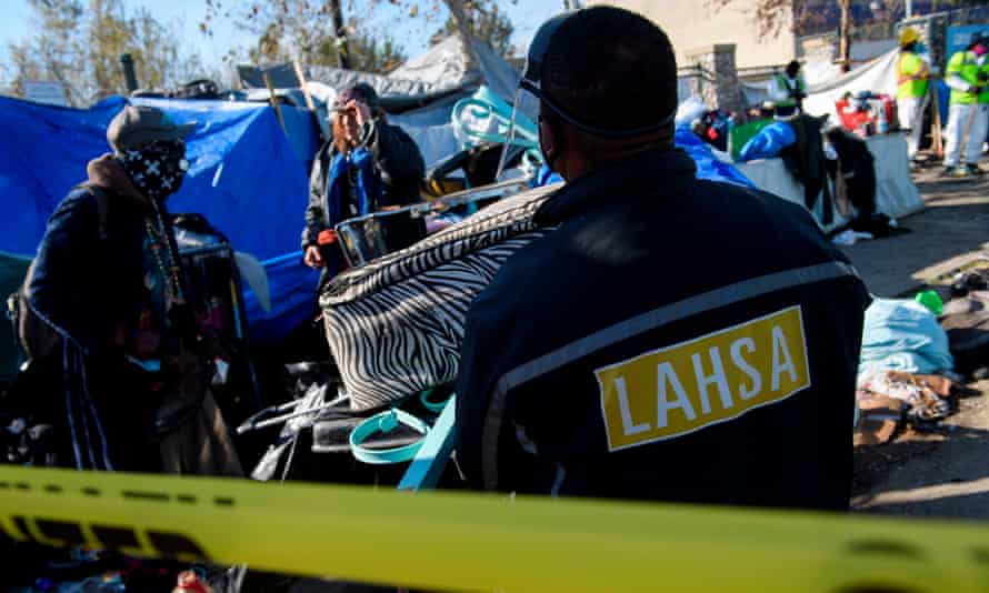 person wearing jacket saying ‘LAHSA’ behind caution tape, near tents