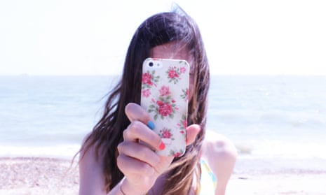 Girl taking photo with her mobile phone, face hidden behind the screen, with the ocean as a backdrop
