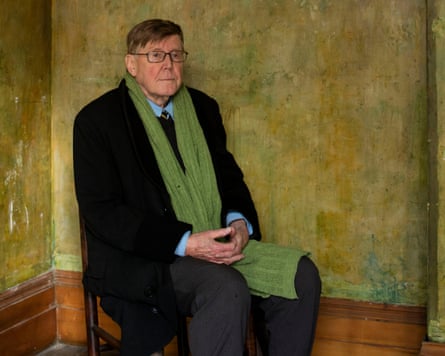 Wearing a black coat and a long green scarf over a blue shirt and tie, he sits on a chair in the corner of a room painted a mottled algae-green