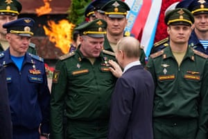 Surrounded by men in military uniforms, Vladimir Putin, facing away from the camera, pats an officer, who is wearing a war hero badge, on the chest