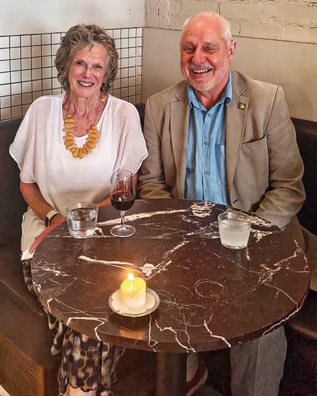 A smiling woman in a white top and a smiling man in a light coloured suit and shirt, sitting at a restaurant table with drinks and a candle on