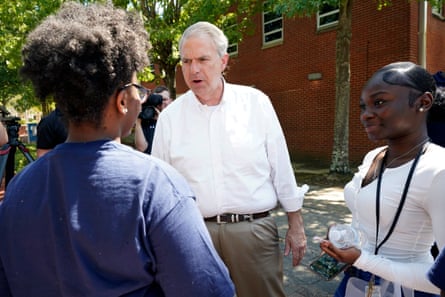 A middle-aged white man in a white collared shirt, no tie, speaks with two young Black women, one wearing a blue T-shirt and the other a long-sleeved white T-shirt.