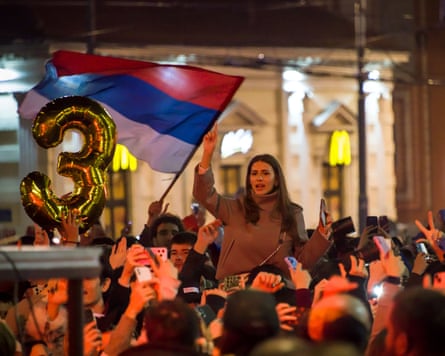 A woman sits on shoulders raising her hand amid a crowd celebrating in Belgrade, with a gold “3” balloon and flag held aloft nearby