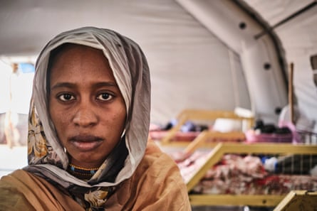 A woman wearing a head scarf looks directly at the camera. Behind her is a row of beds.