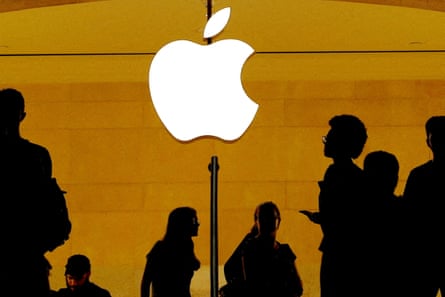 Silhouetted customers walk past a white Apple logo on a light brown backdrop inside an Apple store at Grand Central Station in New York.