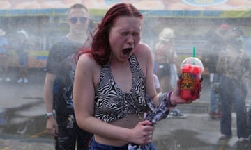 A woman holding a frozen drink gasps with open mouth and scrunched eyes to cold water at a cooling station during a period of hot weather on the midway of the Calgary Stampede in Alberta, with the background obscured by mist. A man stands behind her in the queue and in the background is a Now Mini Donut stand with people in Stetsons mingling in front of it
