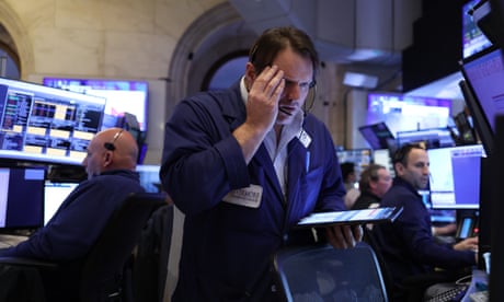 Traders at the New York Stock Exchange: one man holds his hand to his head as he stands looking at a hand-held screen; others sit looking at screens behind him.