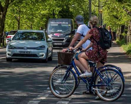 Two cyclists wait for an opening at a T-junction as cars approach under a sunny canopy of trees