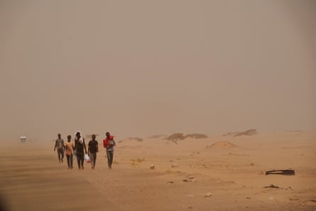 A group of people walk through dust and haze along a road surrounded by sand.
