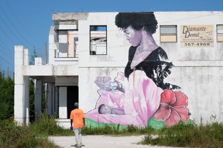 a person in an orange t-shirt walks in front of a white roofless building with a mural of a woman on the side