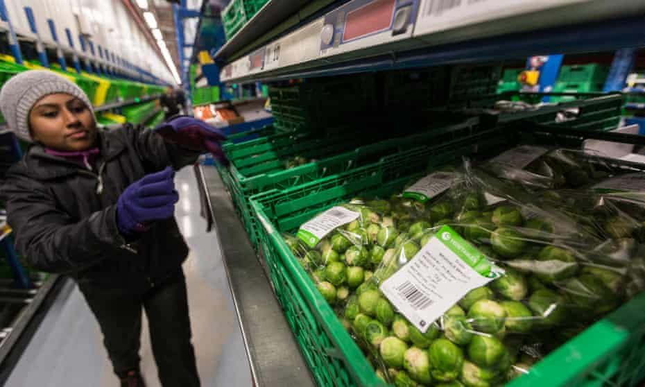 A worker in a wooly hat heading down a narrow aisle towards a hopper full of bags of Brussels sprouts