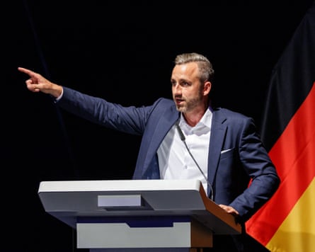 Ulrich Siegmund holds his arm up as he addresses delegates. He is young, with short blond hair, and stands in front of a German flag.