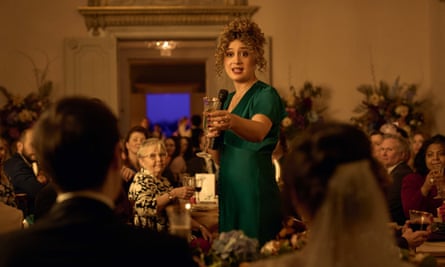 rose matafeo holds a microphone, giving a speech at a wedding dinner, the picture taken from behind bride and groom