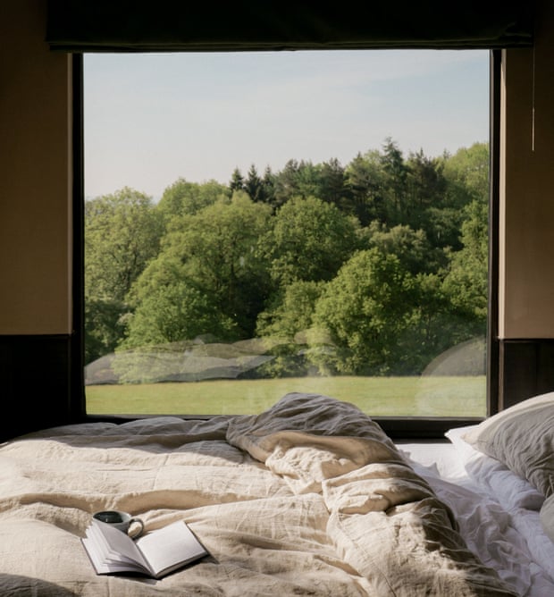A bed with a book and cup of coffee on the sheets, looking out a large window to a lawn and trees