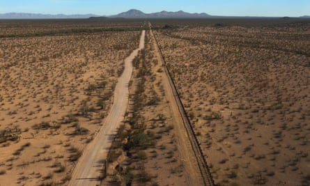 An aerial view of a flat sandy landscape with scrub brush, and a two-lane road and train tracks leading into low mountains far in the distance.