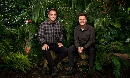 Two men wearing dark clothes, smile into camera, both sitting on a log in the middle of a jungle setting