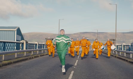 A man walking along a road in a green tracksuit, followed by a group of men in orange tracksuits