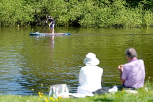 Two people with their backs to the camera sit on the riverbank as a person paddleboards past in Runnymede, Surrey.
