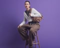 A model with dark hair in a ponytail and red lipstick, sitting on a stool wearing a grey crew neck, a white ballet wrap and brown trousers, and holding a brown bag on her shoulder