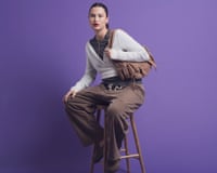 A model with dark hair in a ponytail and red lipstick, sitting on a stool wearing a grey crew neck, a white ballet wrap and brown trousers, and holding a brown bag on her shoulder