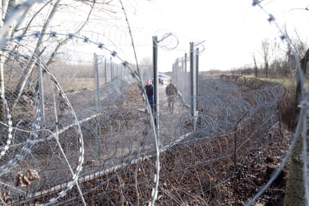 Razor wire in the foreground frames a view of the border fence between Hungary and Serbia, with two guards waling along it