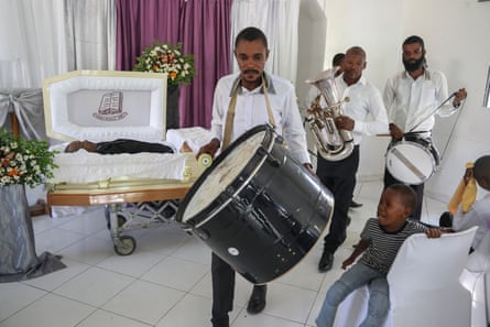 Drummers and English horn player in white dress shirts and black pants parade past a casket on a white-tiled floor while a little boy cries.