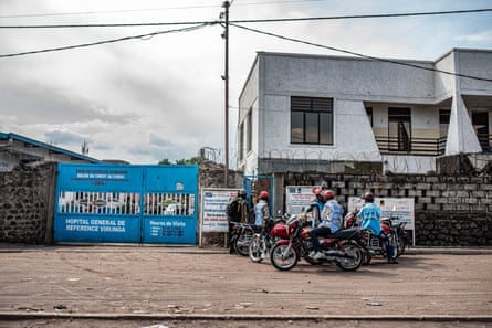 Blue gates are closed in front of a square, low-rise white building. A group of motorbike riders cluster in front of it.
