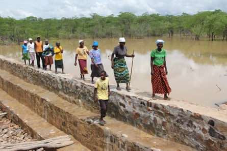 Ten African women and men walking along a stone dam wall with partially submerged acacia-type trees in the background