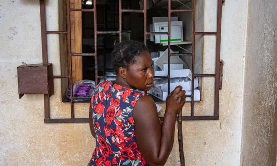 A woman supporting herself with a stick waits by a window to receive medication