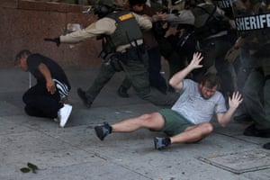 Two men lie on the ground, one with his hands in the air and the other clutching his leg, as riot police move in behind them