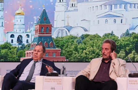 Sergei Lavrov and Errol Musk on stage with a backdrop depicting the Kremlin’s domed buildings