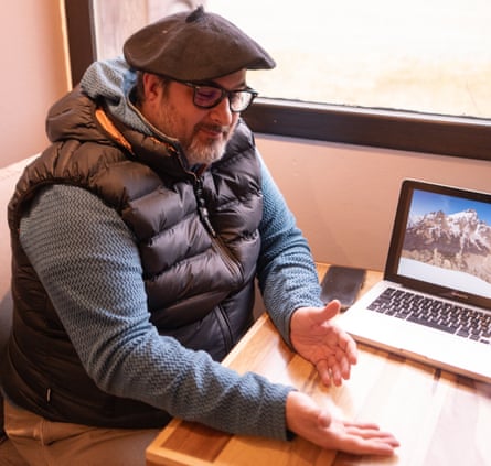 A man wearing a beret, glasses and gilet sits at a desk with a laptop on it.
