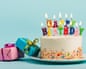 A white cake with colourful "Happy Birthday" candles sits beside wrapped gift boxes on a plate