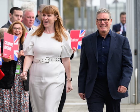Angela Rayner in a white belted dress walks alongside Keir Starmer in a dark suit and glasses