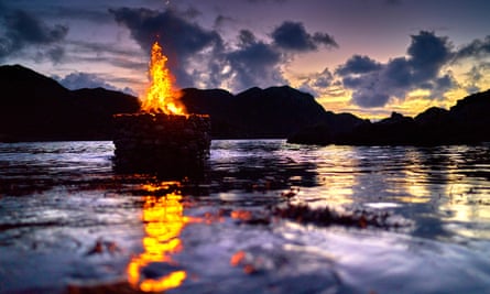 Flames rise from a circle of stones surrounded by water, at sunset